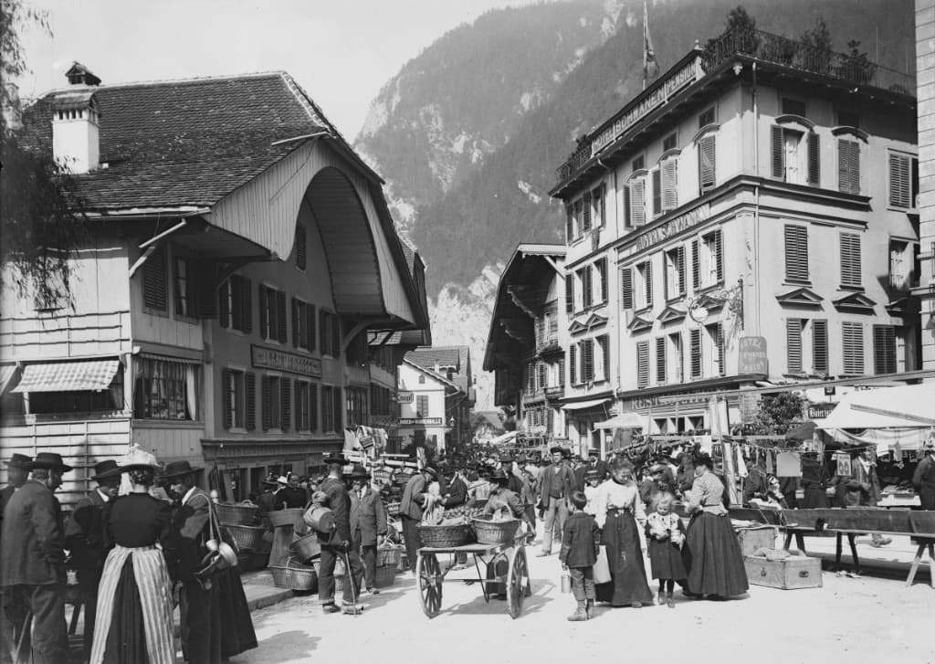 Historische Aufnahme eines belebten Ortsbildes mit traditionellen Holzhäusern und Bergkulisse